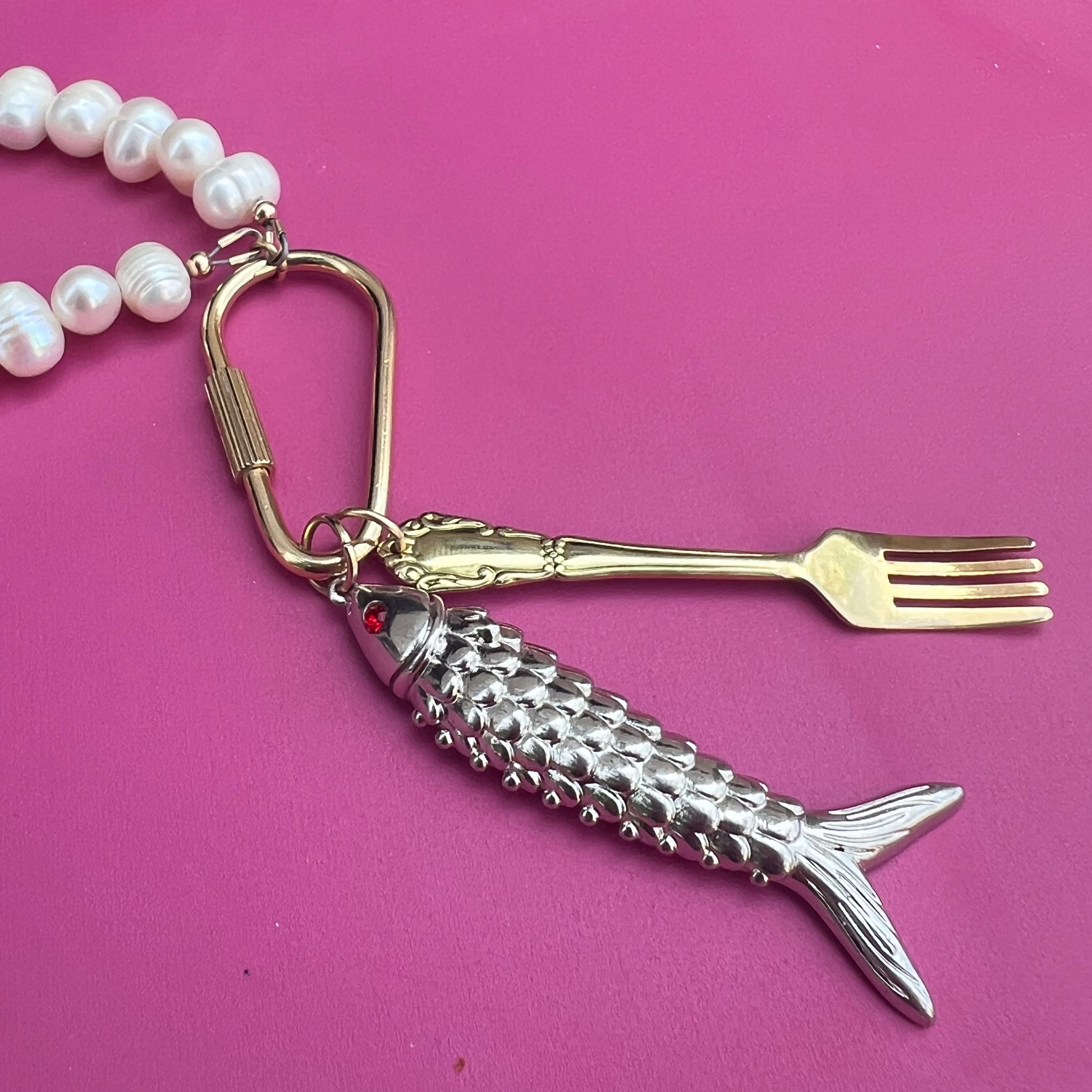 Fish-shaped keychain with fork and pearl necklace on a pink surface