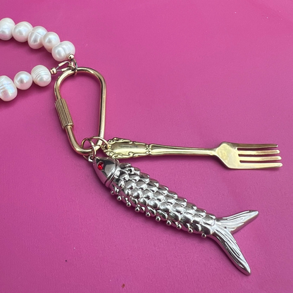 Fish-shaped keychain with fork and pearl necklace on a pink surface